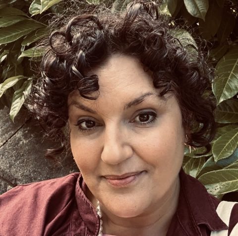 Yasmin A Darling, a woman with dark curly hair, smiling, photographed among green leaves